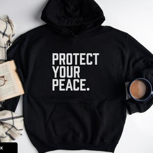 A bold typographic design reading PROTECT YOUR PEACE. Design is white, printed on a Gildan 18500 hoodie in Black colorway. Garment is arranged flat with a scarf, book, glasses and coffee mug.