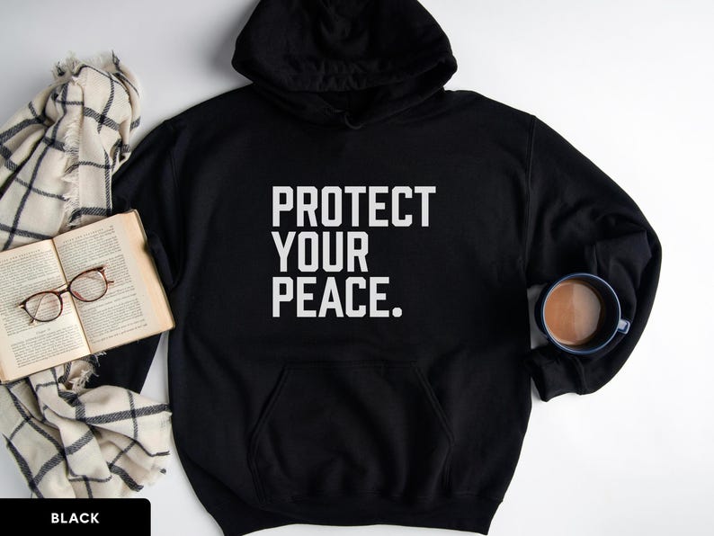 A bold typographic design reading PROTECT YOUR PEACE. Design is white, printed on a Gildan 18500 hoodie in Black colorway. Garment is arranged flat with a scarf, book, glasses and coffee mug.