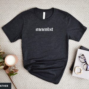 A minimalist typographic design that reads "mnmlst". Design is white on an Dark Heather Grey Bella+Canvas 3001 t-shirt. Garment is displayed flat with two books, stylish glasses, candle and decorative eucalyptus branch.