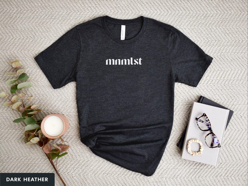 A minimalist typographic design that reads "mnmlst". Design is white on an Dark Heather Grey Bella+Canvas 3001 t-shirt. Garment is displayed flat with two books, stylish glasses, candle and decorative eucalyptus branch.
