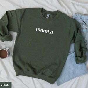 A minimalist typographic design that reads "mnmlst". Design is printed on a Gildan 18000 sweatshirt in Military Green colorway. Garment is arranged flat with light blue jeans, glasses and coffee mug.