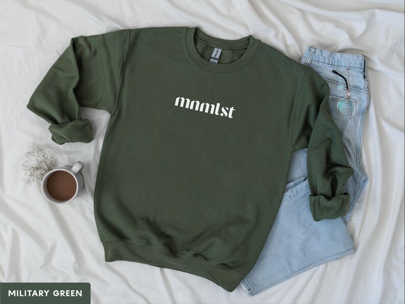 A minimalist typographic design that reads "mnmlst". Design is printed on a Gildan 18000 sweatshirt in Military Green colorway. Garment is arranged flat with light blue jeans, glasses and coffee mug.