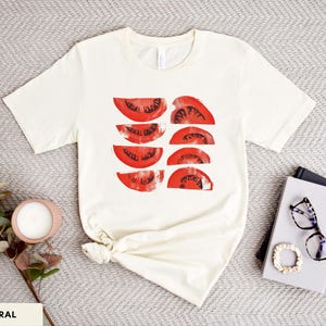 A beautiful, rustic print of eight red ripe tomato slices. Design is multi-color on a Natural Bella+Canvas 3001 t-shirt. Garment is displayed flat with two books, stylish glasses, candle and decorative eucalyptus branch.
