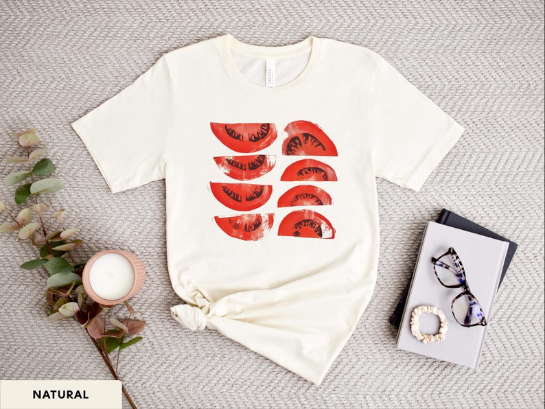 A beautiful, rustic print of eight red ripe tomato slices. Design is multi-color on a Natural Bella+Canvas 3001 t-shirt. Garment is displayed flat with two books, stylish glasses, candle and decorative eucalyptus branch.