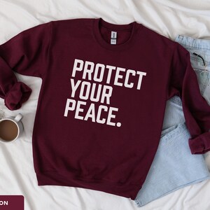 A bold typographic design reading PROTECT YOUR PEACE. Design is white, printed on a Gildan 18000 sweatshirt in Maroon colorway. Garment is arranged flat with light blue jeans, glasses and coffee mug.