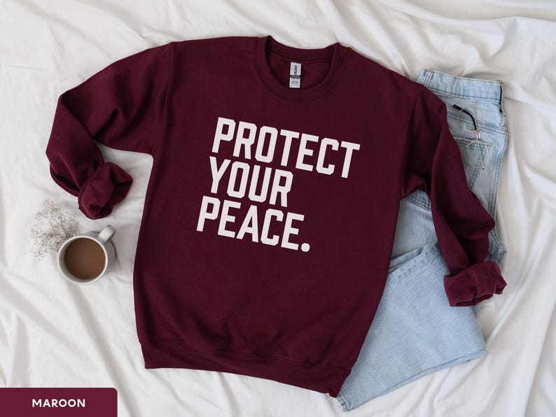 A bold typographic design reading PROTECT YOUR PEACE. Design is white, printed on a Gildan 18000 sweatshirt in Maroon colorway. Garment is arranged flat with light blue jeans, glasses and coffee mug.