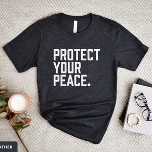 A bold typographic design reading PROTECT YOUR PEACE. Design is white on an Dark Heather Bella+Canvas 3001 t-shirt. Garment is displayed flat with jeans, two books, candle and decorative eucalyptus branch.
