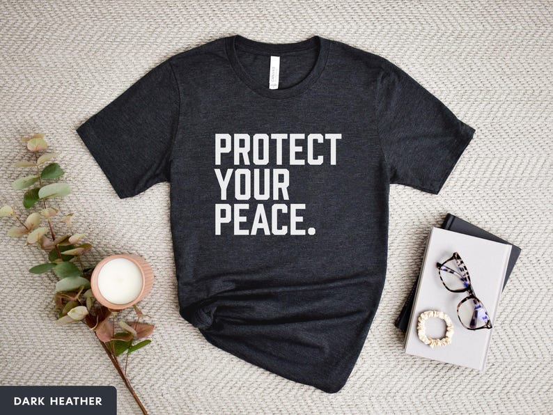 A bold typographic design reading PROTECT YOUR PEACE. Design is white on an Dark Heather Bella+Canvas 3001 t-shirt. Garment is displayed flat with jeans, two books, candle and decorative eucalyptus branch.