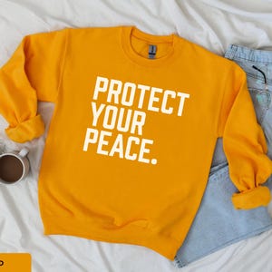 A bold typographic design reading PROTECT YOUR PEACE. Design is white, printed on a Gildan 18000 sweatshirt in Gold colorway. Garment is arranged flat with light blue jeans, glasses and coffee mug.