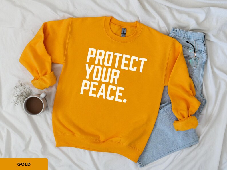 A bold typographic design reading PROTECT YOUR PEACE. Design is white, printed on a Gildan 18000 sweatshirt in Gold colorway. Garment is arranged flat with light blue jeans, glasses and coffee mug.