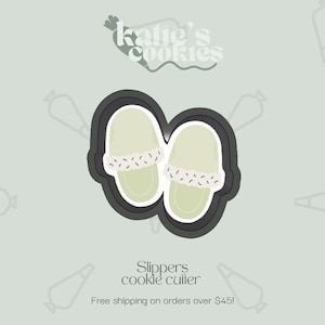 May include: A cookie cutter in the shape of a pair of slippers. The slippers are light green with a white trim and a black outline. The text "Slippers cookie cutter" is below the image.