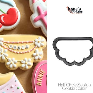 May include: A yellow cookie decorated with white daisy flowers and the name "ANNIE" in bead letters. The cookie is surrounded by other decorated cookies, including a cherry and a pink cross. A black half-circle scallop cookie cutter is also shown.
