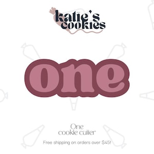 May include: A pink cookie cutter in the shape of the word "one". The cookie cutter is on a white background with the text "katie's cookies" above it.
