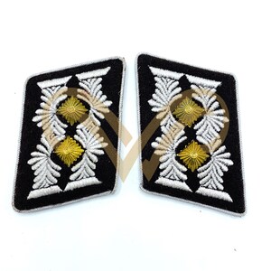 Puede incluir: Par de charreteras negras con intrincados bordados blancos y dorados. El diseño presenta patrones florales estilizados y emblemas centrales dorados, bordeados con ribete plateado. Estos parches de estilo militar son probablemente para un uniforme.