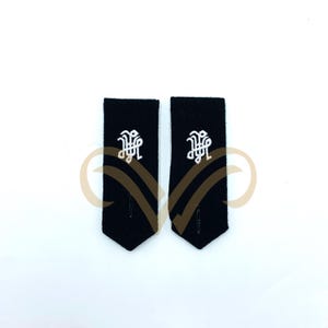 May include: A pair of black shoulder boards, each featuring a white embroidered monogram. The boards are rectangular with a pointed lower edge. The white monogram is an elaborate design, centrally placed. The background is white.
