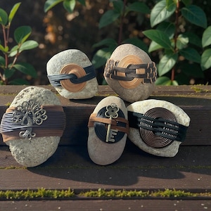May include: Five decorative stones with various designs. Each stone is adorned with a band of black, brown, or tan material, some featuring metal key or tree of life accents. The stones are arranged on a wooden surface.