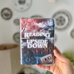 May include: Clear phone case with a graphic design featuring the text "READING THE UPSIDE DOWN." The design includes a silhouette of a person on a bicycle, a cassette tape, and a stormy sky with lightning.