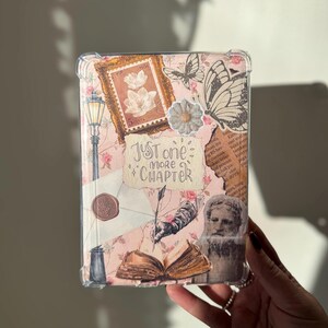 May include: A pink and white book cover with a clear protective case. The cover features a variety of vintage-style images, including a butterfly, a stamp, a quill pen, and a book. The text "Just one more chapter" is written in black cursive lettering.