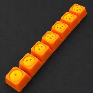 Pode incluir: Um conjunto de sete teclas quadradas laranja para teclado mecânico dispostas numa linha diagonal. Cada tecla apresenta um centro circular amarelo com um número variável de estrelas vermelhas, de uma a sete, em relevo no topo.