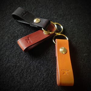 Könnte beinhalten: Drei Leder-Schlüsselanhänger mit Messing-Schlüsselringen und Schraubverschlüssen. Die Schlüsselanhänger sind braun, schwarz und orange.