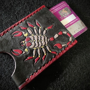 Pode incluir: Porta-cartões de couro preto com um design de escorpião vermelho e amarelo. As bordas são vermelhas com costuras vermelhas. Um cartão roxo é parcialmente visível dentro. O texto "AUTHORISED SIGNA" é visível no cartão.