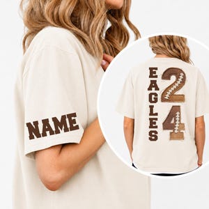 May include: A beige short-sleeve t-shirt featuring the word NAME in brown block letters on the sleeve. The back of the shirt displays the word EAGLES vertically in brown, alongside the number 24 with a brown football-textured applique design.