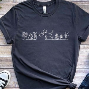 May include: Dark gray t-shirt featuring a white line drawing of a dog watering potted plants. The design includes various plants and a dog holding a watering can. The shirt has a crew neck and short sleeves.