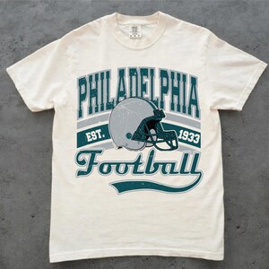 Może przedstawiać: Kremowy t-shirt z kr&oacute;tkim rękawem z nadrukiem graficznym zawierającym napis PHILADELPHIA EST. 1933 Football. Wz&oacute;r przedstawia srebrny kask do futbolu amerykańskiego z ciemno-turkusową maską ochronną w stylu vintage z efektem przetarcia.