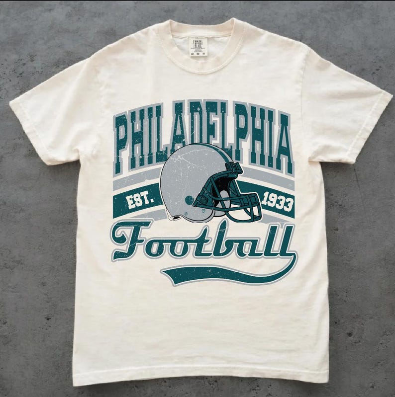 Może przedstawiać: Kremowy t-shirt z kr&oacute;tkim rękawem z nadrukiem graficznym zawierającym napis PHILADELPHIA EST. 1933 Football. Wz&oacute;r przedstawia srebrny kask do futbolu amerykańskiego z ciemno-turkusową maską ochronną w stylu vintage z efektem przetarcia.