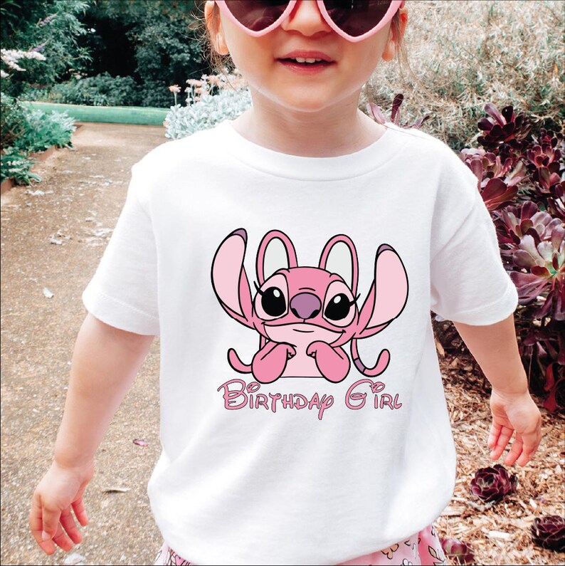 A young girl wearing a white t-shirt with a pink cartoon character and the text "Birthday Girl" printed on it.