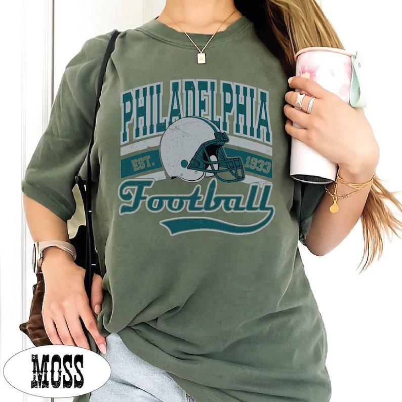 Może przedstawiać: Oliwkowozielony t-shirt z okrągłym dekoltem i nadrukiem graficznym z napisem Philadelphia Football Est. 1933. Wz&oacute;r zawiera biały kask do futbolu amerykańskiego z turkusową maską na twarz na tle przypominającym baner.
