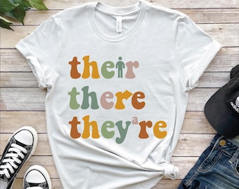 May include: White t-shirt with the words "their, there, they're" in retro-style lettering. The word "their" includes a small stick figure. Black and white sneakers and a black cap are visible.