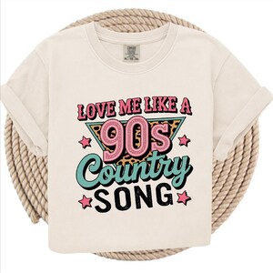 May include: Cream-colored t-shirt with the text "LOVE ME LIKE A 90s COUNTRY SONG" in pink, teal, and black. The design includes stars and a leopard print triangle. The shirt has rolled-up sleeves.