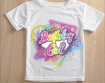 May include: White t-shirt with a colorful "Birthday Girl" graphic. The design features a heart, triangle, and splatter effect in neon colors, including pink, blue, yellow, and purple. The shirt is made of a soft material.