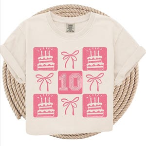 May include: Cream-colored t-shirt with a pink square pattern. The design includes birthday cakes with candles, bows, and the number "10". The shirt is made of a soft material and has rolled-up sleeves.