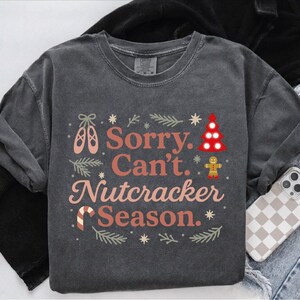 May include: Dark gray long-sleeve shirt with the text "Sorry. Can't. Nutcracker Season." in coral and white. The design includes ballet shoes, a Christmas tree, a gingerbread man, and snowflakes. The shirt is folded with a phone case and jeans in the background.