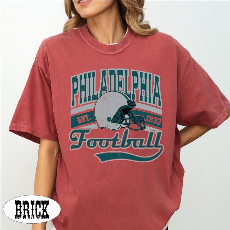 Może przedstawiać: Ceglastoczerwony t-shirt z kr&oacute;tkim rękawem z nadrukiem graficznym zawierającym napis PHILADELPHIA EST. 1933 Football oraz ilustrację białego kasku do futbolu amerykańskiego z ciemnomorską maską ochronną. Wz&oacute;r wykorzystuje ciemnomorskie litery.