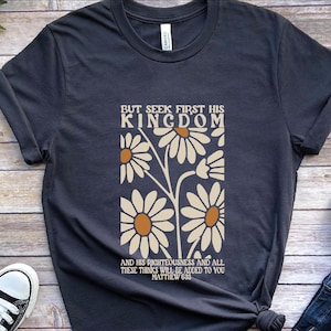 May include: Dark navy blue t-shirt with a daisy floral design and the text "BUT SEEK FIRST HIS KINGDOM AND HIS RIGHTEOUSNESS AND ALL THESE THINGS WILL BE ADDED TO YOU MATTHEW 6:33". The shirt is a casual style.