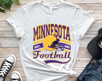 May include: White t-shirt featuring a distressed design with the text "MINNESOTA Football" in purple and yellow. The design includes a football helmet and the year "1961". The shirt is paired with black sneakers and a black cap.