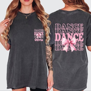 May include: Dark gray t-shirt with the word "DANCE" in pink, repeated vertically, and a pink bow graphic on the back. The front of the shirt has the word "DANCE" repeated vertically with the name "Olivia" below.