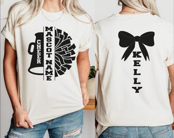 May include: Cream-colored t-shirt with a black cheerleading design. The front features a megaphone, pom-pom, and the words "CHEER" and "MASCOT NAME". The back displays a large bow and the name "KELLY".