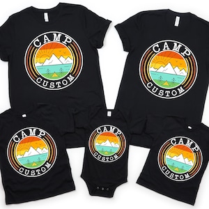 May include: Black t-shirts and a onesie with a circular graphic. The graphic features a mountain range, sun, and lake scene with the words "CAMP CUSTOM" in a circular design. The shirts are short-sleeved and the onesie has short sleeves.