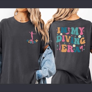 May include: Dark grey t-shirt with a colorful graphic on the front and back. The front features a surfer graphic with the word "CUSTOM". The back of the shirt says "IN MY DIVING ERA" in a retro font with floral accents.