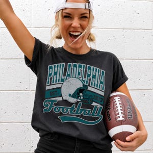Może przedstawiać: Ciemnoszary t-shirt z kr&oacute;tkim rękawem z postarzanym nadrukiem graficznym z napisem Philadelphia Football Est. 1933 oraz ilustracją kasku do futbolu amerykańskiego pośrodku. Wz&oacute;r wykorzystuje turkusowe i białe kolory na ciemnej tkaninie.