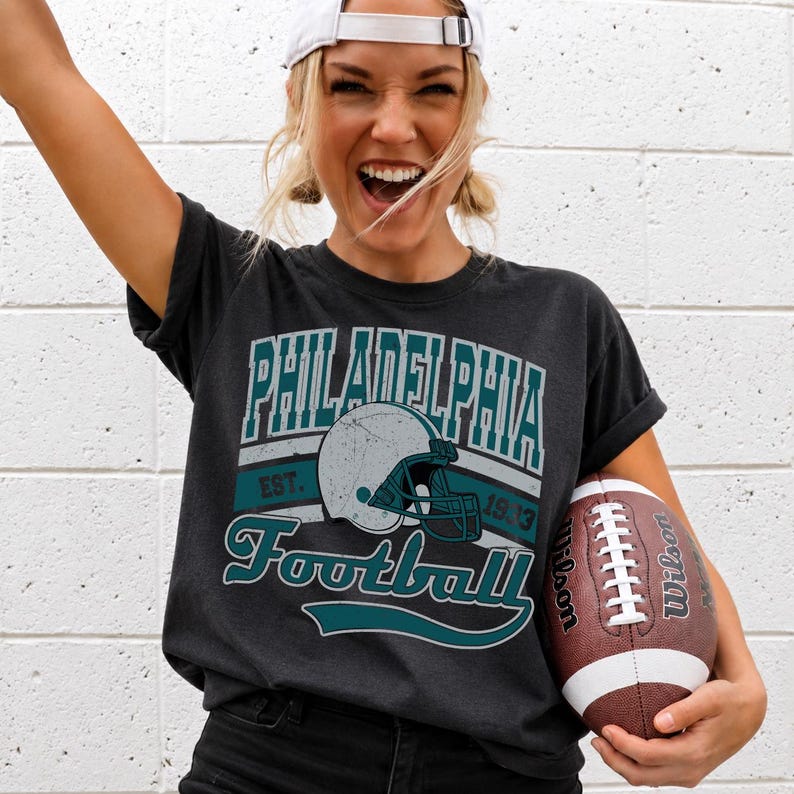 Może przedstawiać: Ciemnoszary t-shirt z kr&oacute;tkim rękawem z postarzanym nadrukiem graficznym z napisem Philadelphia Football Est. 1933 oraz ilustracją kasku do futbolu amerykańskiego pośrodku. Wz&oacute;r wykorzystuje turkusowe i białe kolory na ciemnej tkaninie.