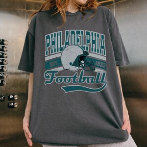 Może przedstawiać: Ciemnoszary t-shirt z kr&oacute;tkim rękawem z nadrukiem graficznym zawierającym napis Philadelphia Football Est. 1933. Wz&oacute;r przedstawia biały kask do futbolu amerykańskiego z turkusową maską, otoczony turkusowymi napisami i liniami dekoracyjnymi.