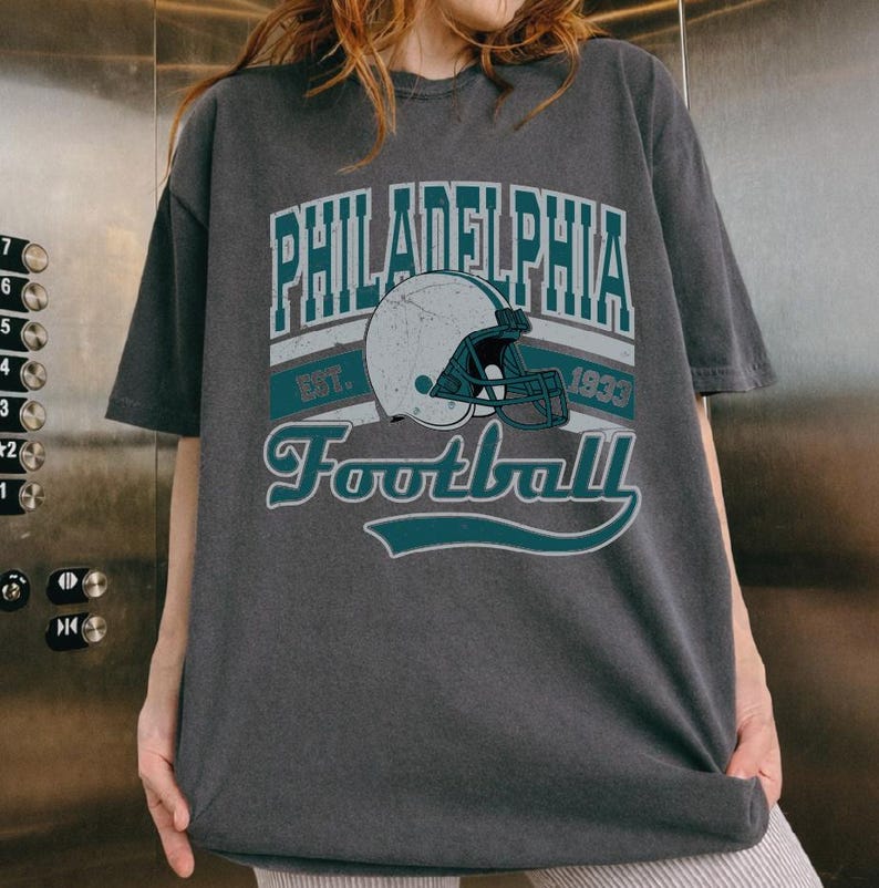 Może przedstawiać: Ciemnoszary t-shirt z kr&oacute;tkim rękawem z nadrukiem graficznym zawierającym napis Philadelphia Football Est. 1933. Wz&oacute;r przedstawia biały kask do futbolu amerykańskiego z turkusową maską, otoczony turkusowymi napisami i liniami dekoracyjnymi.