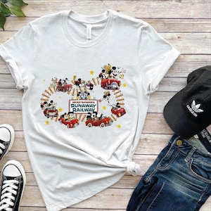 May include: White t-shirt featuring a colorful graphic of Mickey and Minnie Mouse in red cars on a railway track. The text "Mickey & Minnie's Runaway Railway" is displayed on a sign. A black Adidas cap and blue jeans are also visible.