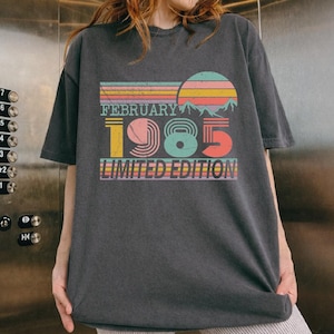 May include: Dark gray t-shirt with a retro design featuring the text "FEBRUARY 1985 LIMITED EDITION." The design includes a colorful sunset, mountains, and the numbers 1985 in a vintage font. The shirt is a relaxed fit.