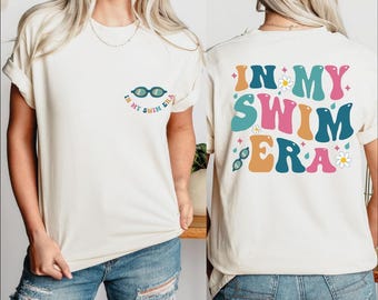 May include: Cream-colored t-shirt with the phrase "IN MY SWIM ERA" in colorful, retro-style lettering. The front features a small graphic of goggles and text. The back of the shirt has a larger design with the same text and additional graphics.